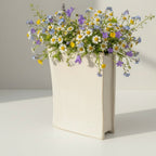 A white vase filled with a vibrant bouquet of colorful flowers, including daisies, chrysanthemums, and other blooms, placed on a white surface.
