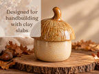A ceramic acorn-shaped container with a textured surface sits on a wooden surface, surrounded by fallen autumn leaves.