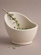 A white ceramic bowl with a curved shape, containing a sprig of green herbs or a similar plant.