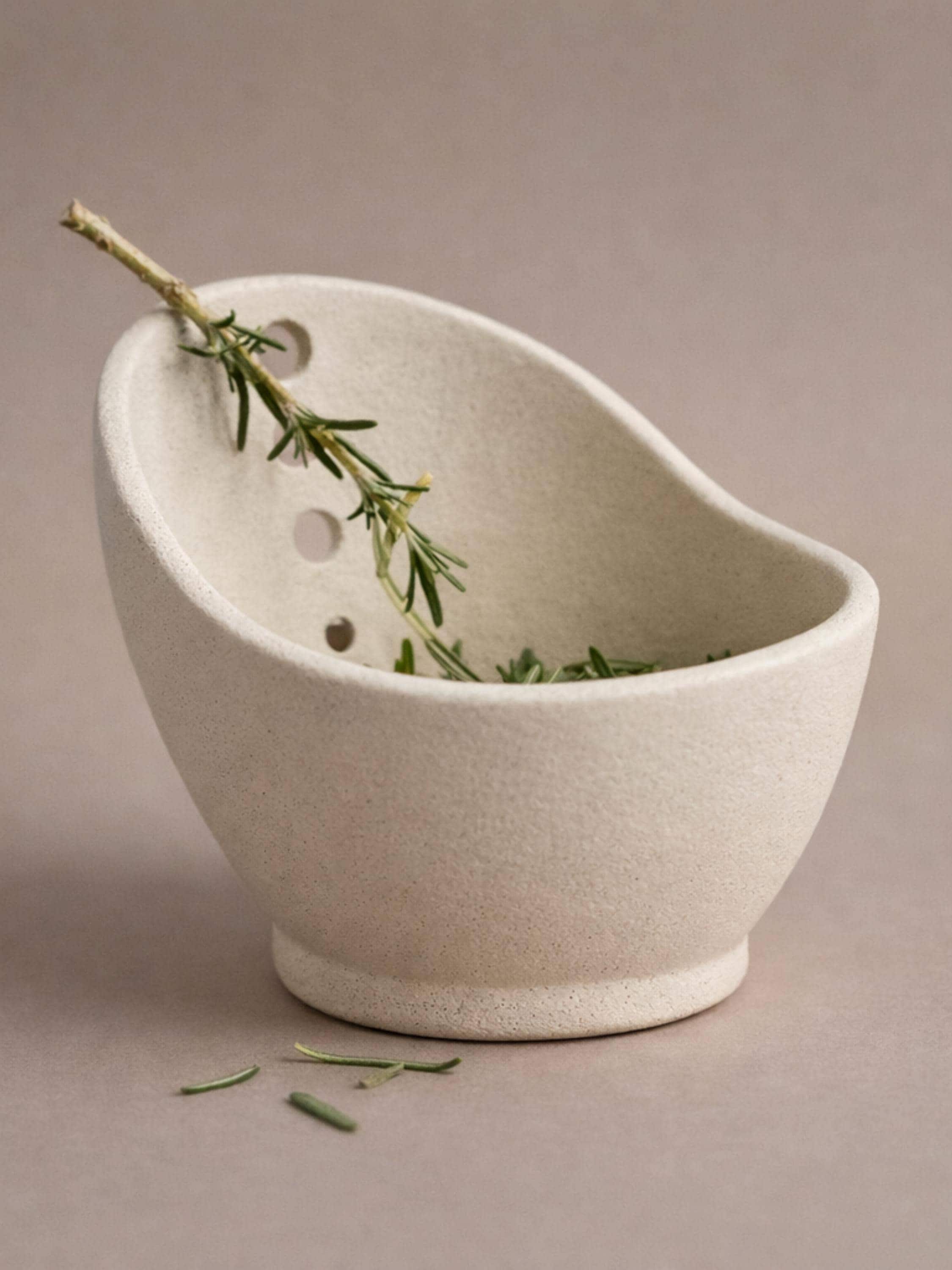 A white ceramic bowl with a curved shape, containing a sprig of green herbs or a similar plant.