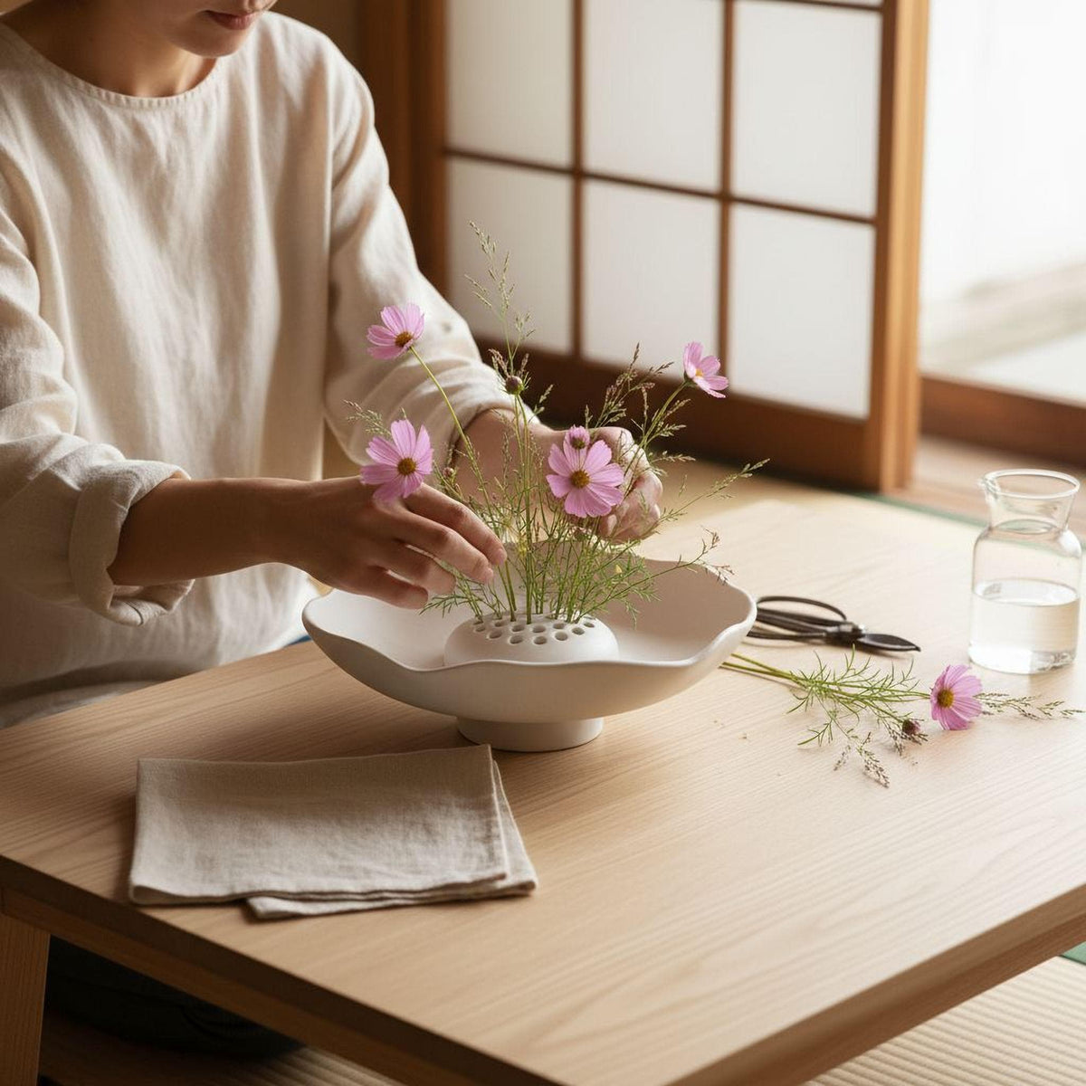 A person is arranging pink flowers in a white bowl on a wooden table, with a pair of scissors nearby.