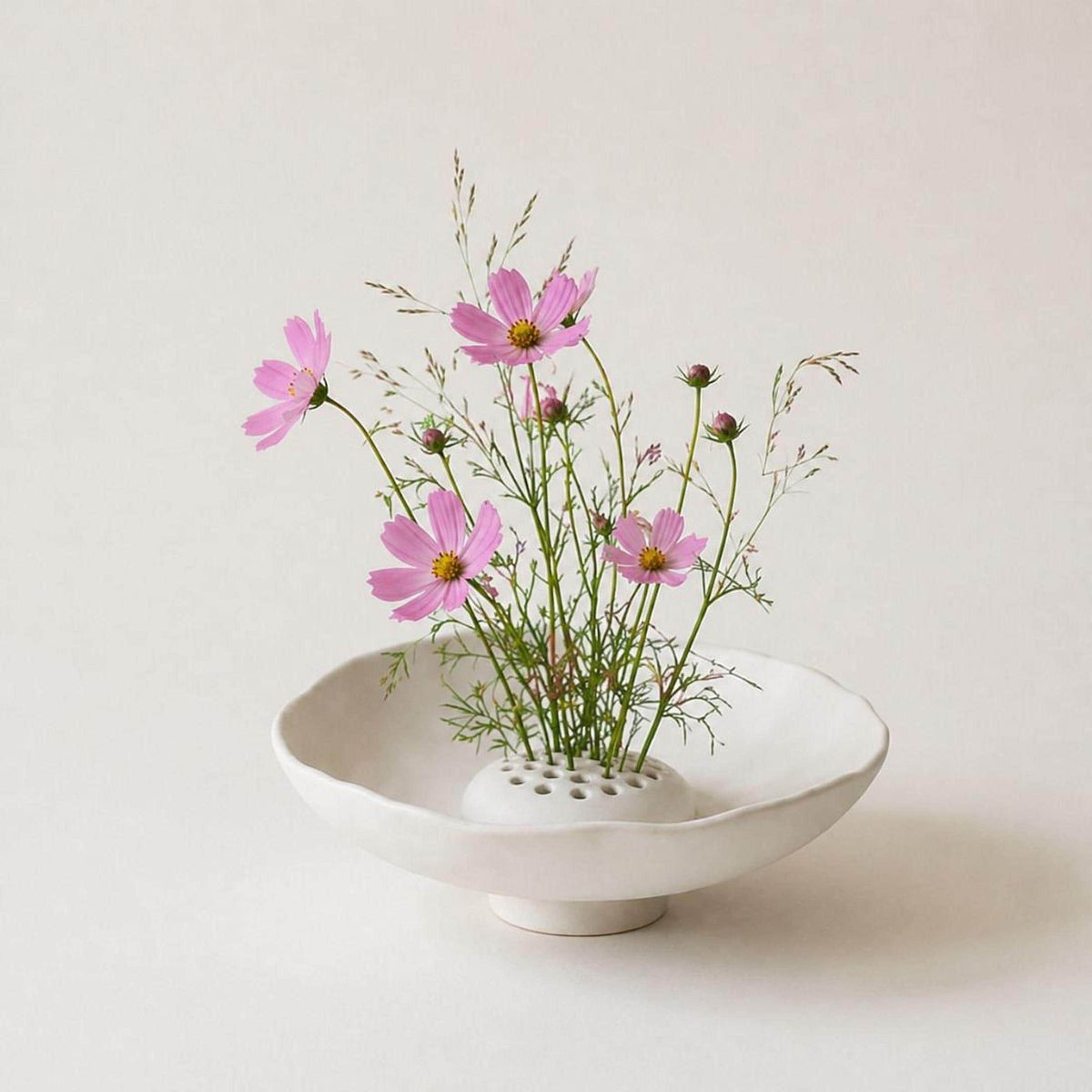 A white ceramic bowl with a floral arrangement of pink flowers and green foliage placed inside it, set against a plain white background.