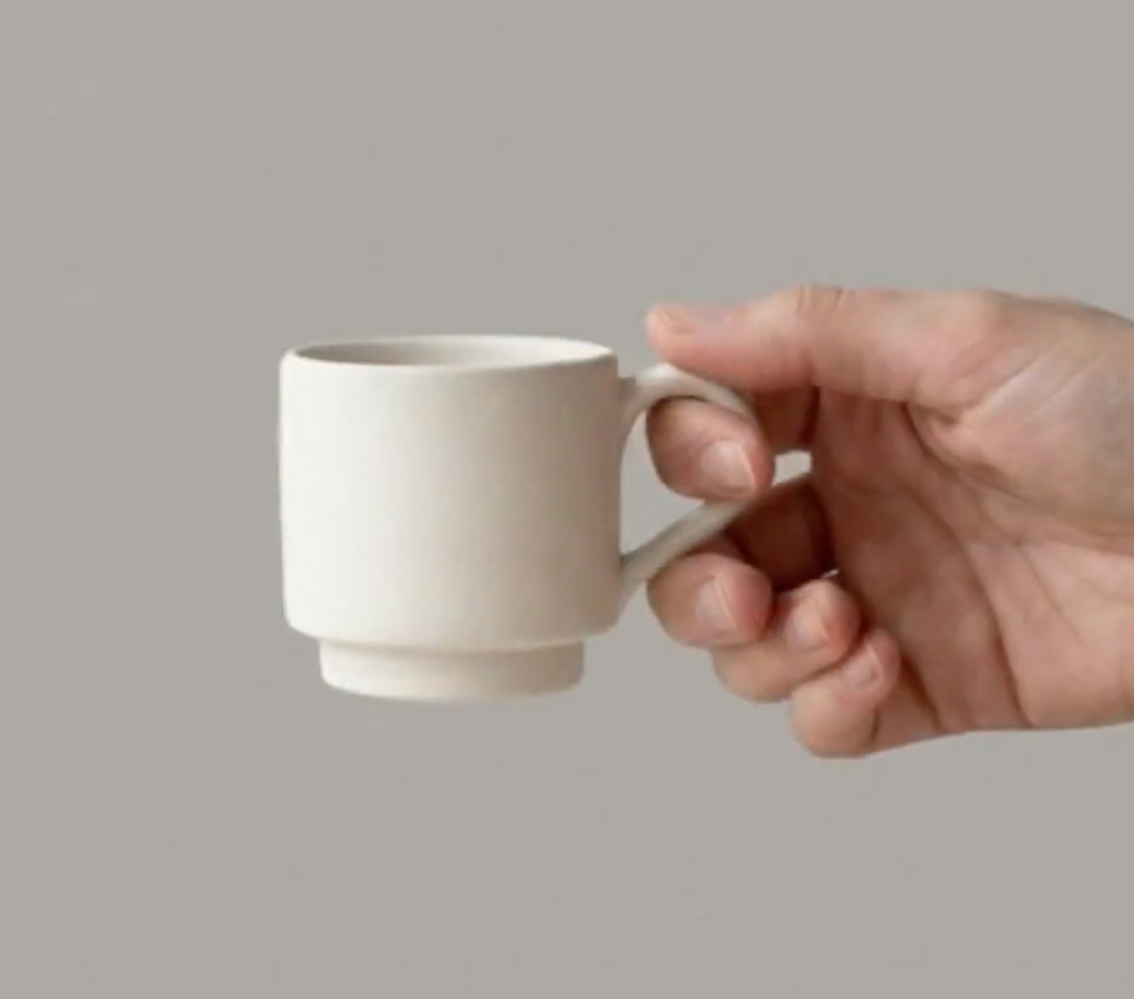 A hand holding a white ceramic mug against a gray background.