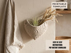 a heart-shaped planter with a plant inside, mounted on a wall. The planter has a decorative design and appears to be made of a light-colored material. There is a towel hanging on the wall next to the planter.
