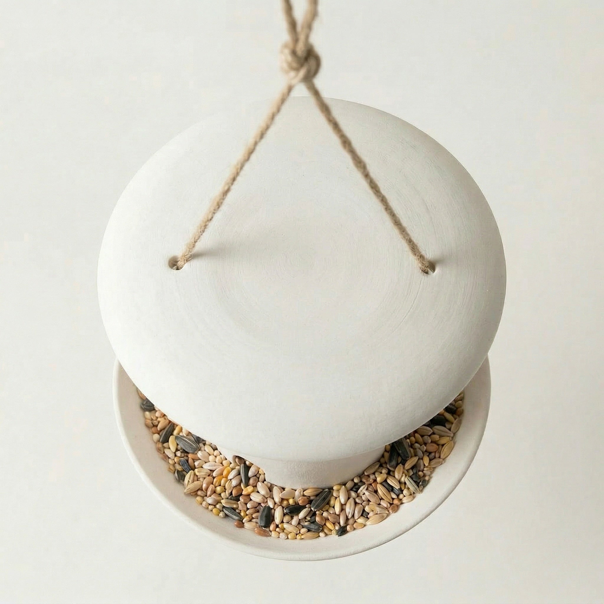 A white ceramic bird feeder with a hanging rope and a bowl filled with birdseed.