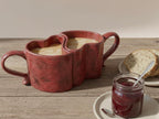 A rustic wooden table with two heart-shaped red mugs filled with a warm beverage, accompanied by a jar of jam and a slice of bread.