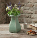 A green ceramic vase filled with purple and white flowers sits on a wooden table, accompanied by a cup of tea, a plate of nuts, and a book.