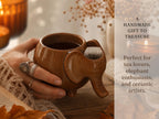 A hand holding a brown ceramic mug with an elephant-shaped design, with a candle and other decorative items visible in the background.
