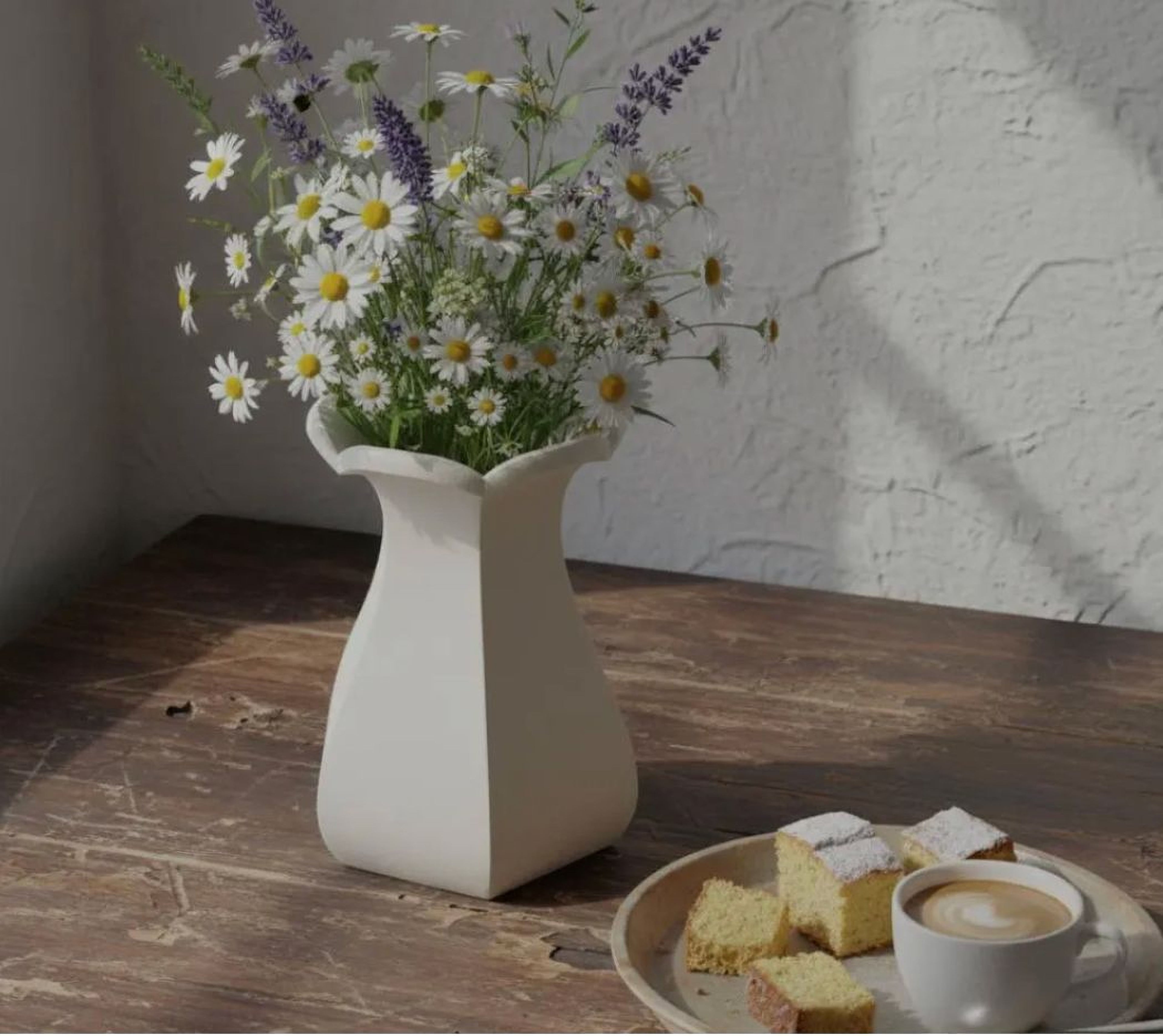 A white vase filled with a bouquet of white and purple flowers sits on a wooden table, accompanied by a plate of pastries and a cup of coffee.