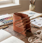 a wooden table with various art supplies, including a ceramic container with multiple paintbrushes, a sketchbook, and a jar filled with art materials.