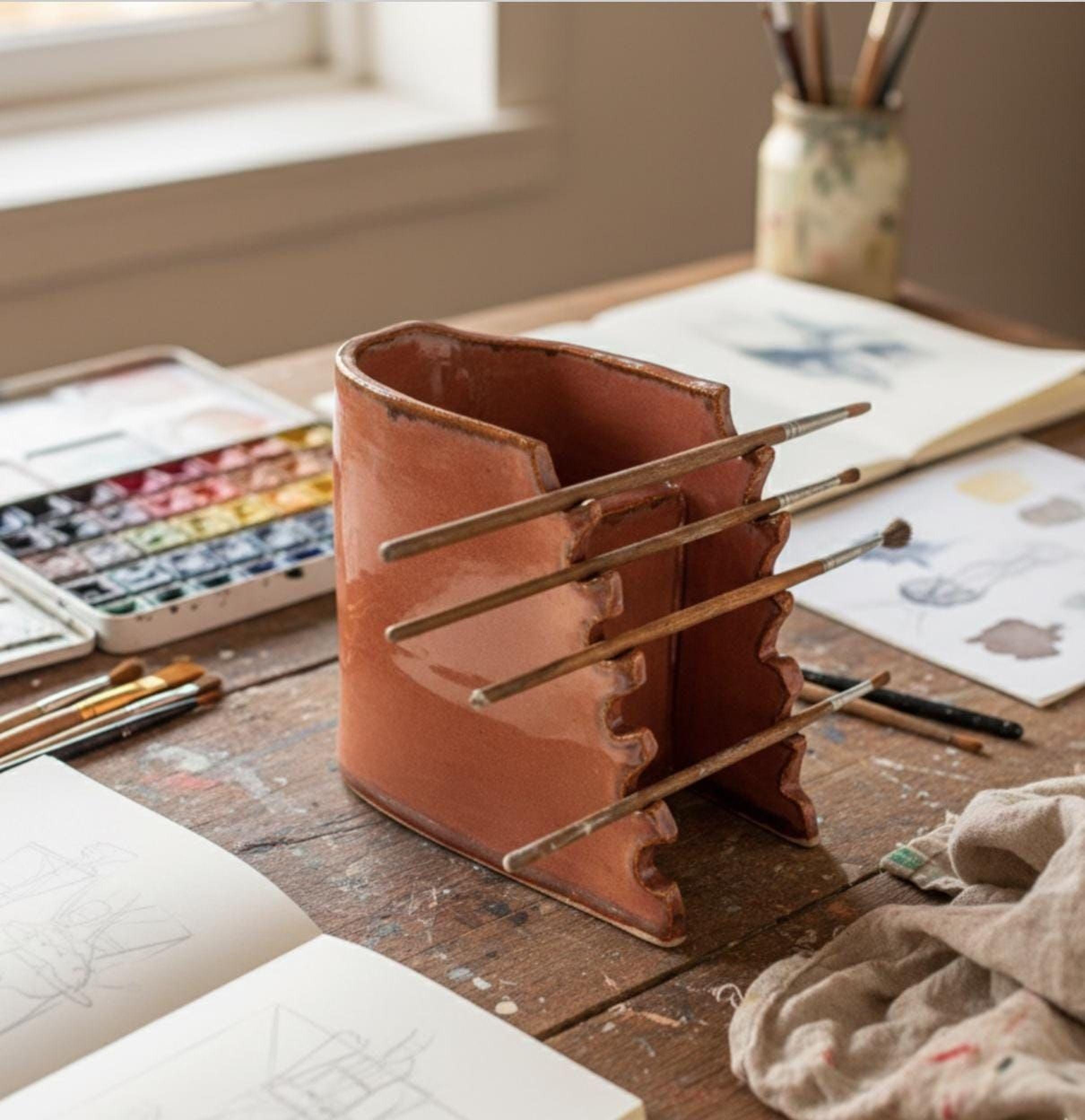 a wooden table with various art supplies, including a ceramic container with multiple paintbrushes, a sketchbook, and a jar filled with art materials.