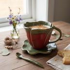 A red ceramic mug filled with a warm beverage sits on a wooden table, accompanied by a green spoon, a small vase of colorful wildflowers, and a partially eaten cookie.