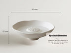 a white ceramic bowl with a hole in the center, placed on a white surface. The bowl appears to be empty, and there is a line indicating the dimensions of the bowl.