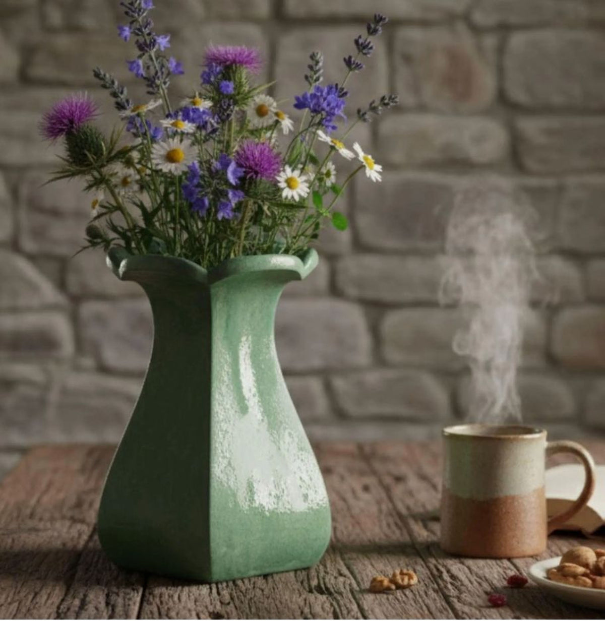 A green vase filled with purple and white flowers sits on a wooden table, with a steaming mug of coffee nearby.