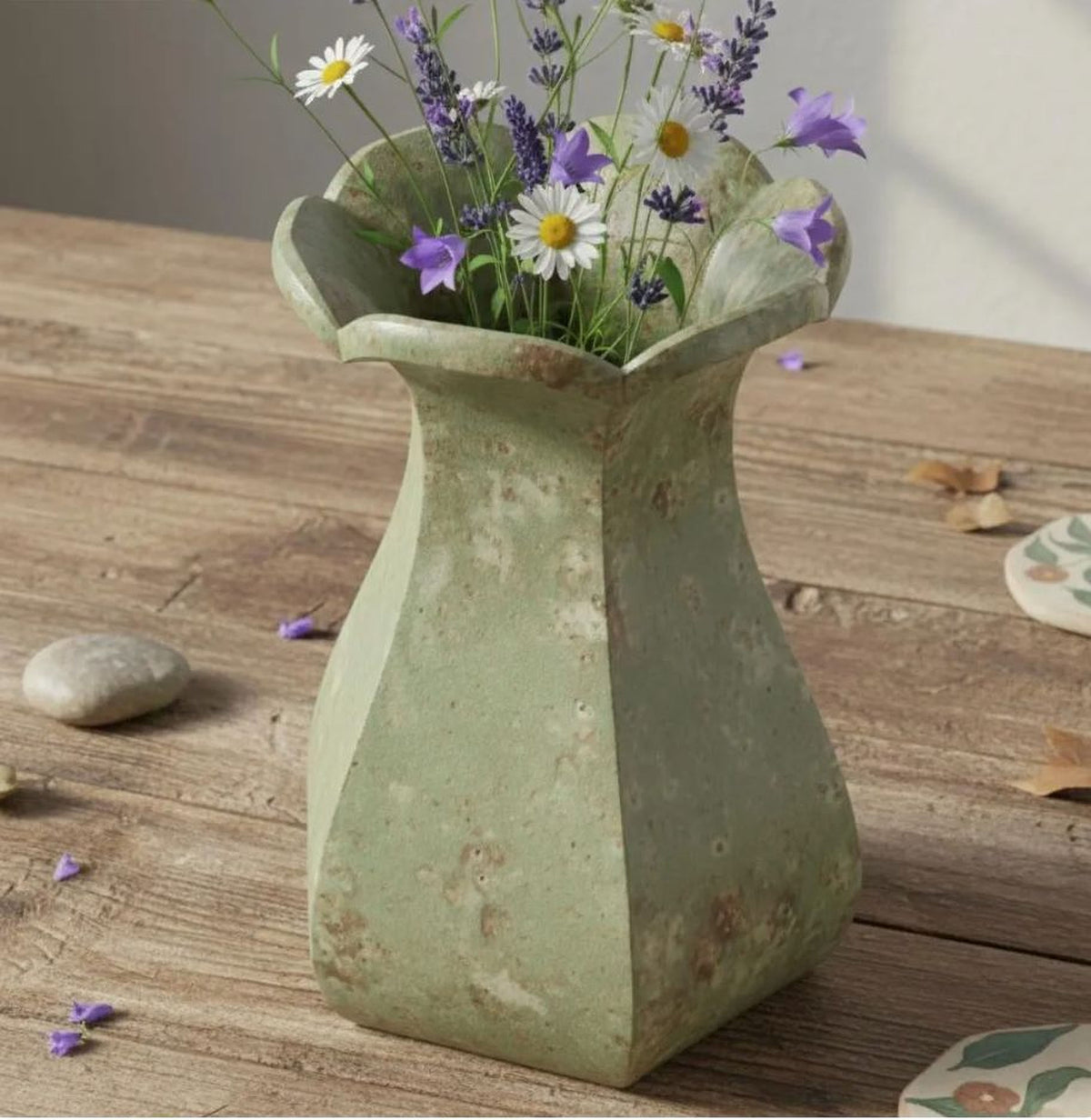 A green vase filled with purple and white flowers sits on a wooden surface, surrounded by scattered petals.
