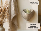 a heart-shaped planter with a plant growing in it, placed on a wall next to a beige shawl and a vase of dried flowers.