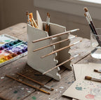 a wooden table with various art supplies, including a white ceramic container holding multiple paintbrushes, a palette of watercolor paints, and some papers with paint splatters.
