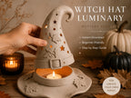 A hand is holding a white ceramic witch's hat with a lit candle inside, surrounded by autumn leaves and pumpkins on a wooden surface.